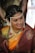 A woman dressed in traditional Indian attire with intricate jewelry, including a pearl headdress and a nose ring. The sari is a combination of rich maroon and vibrant orange with gold embroidery. Her hair is adorned with white flowers, and she displays a warm smile.