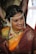 A woman dressed in traditional Indian attire with intricate jewelry, including a pearl headdress and a nose ring. The sari is a combination of rich maroon and vibrant orange with gold embroidery. Her hair is adorned with white flowers, and she displays a warm smile.