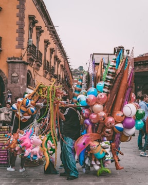 In a bustling street market, vendors hold colorful balloons, inflatable toys, and various decorations. The background features a historic building with arched windows and intricate balconies. People are seen walking and interacting, adding liveliness to the scene.