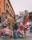 In a bustling street market, vendors hold colorful balloons, inflatable toys, and various decorations. The background features a historic building with arched windows and intricate balconies. People are seen walking and interacting, adding liveliness to the scene.