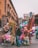 In a bustling street market, vendors hold colorful balloons, inflatable toys, and various decorations. The background features a historic building with arched windows and intricate balconies. People are seen walking and interacting, adding liveliness to the scene.