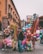 In a bustling street market, vendors hold colorful balloons, inflatable toys, and various decorations. The background features a historic building with arched windows and intricate balconies. People are seen walking and interacting, adding liveliness to the scene.