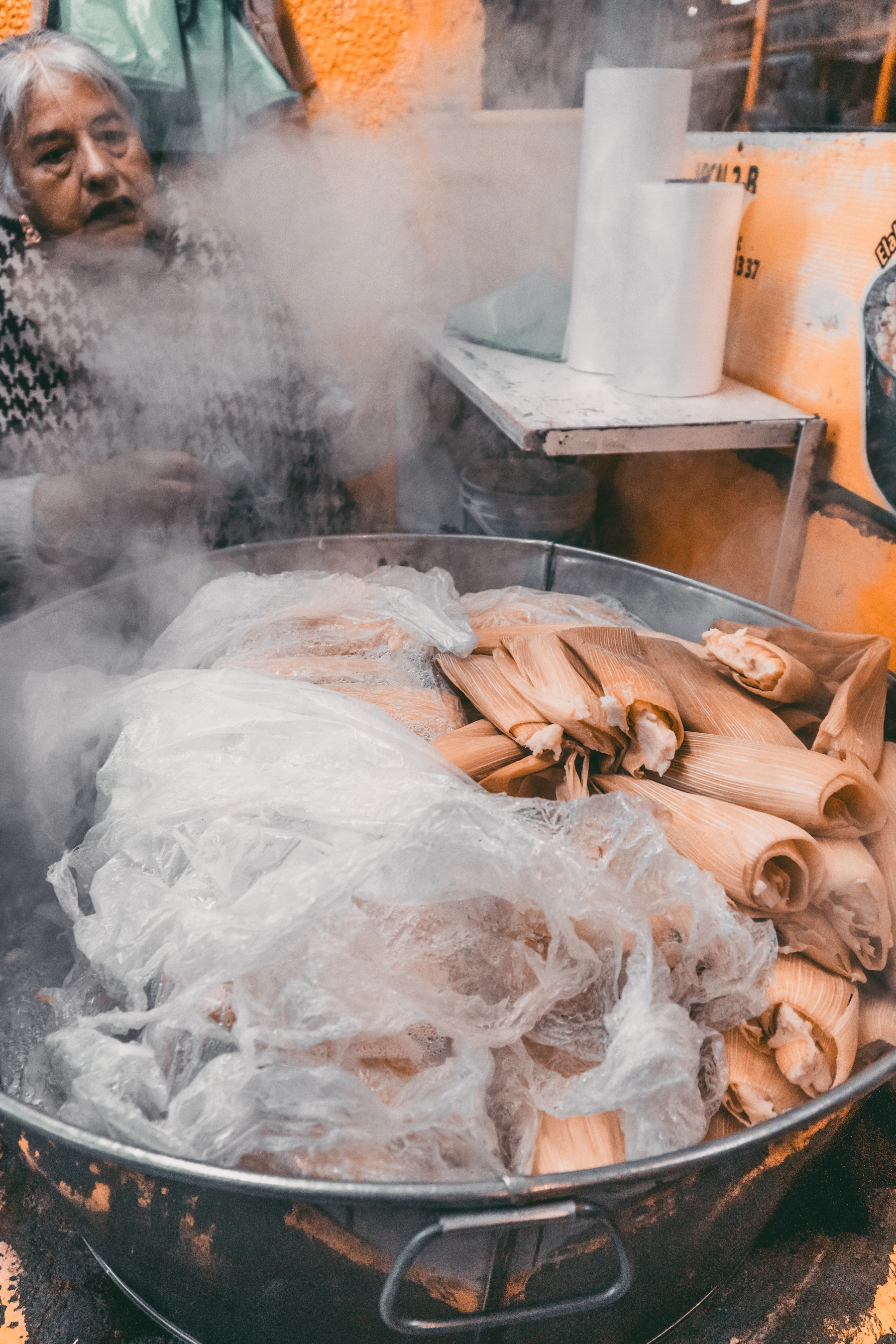 Tamales wrapped in corn husks heaped in a big steaming pot, a cook behind the steam
