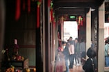 A corridor in a traditional or religious setting with red decorations hanging from the ceiling. People are gathered, some holding incense sticks. An offering table with fruits and candles is visible on the left. The atmosphere is calm, with a focus on prayer or ritual.