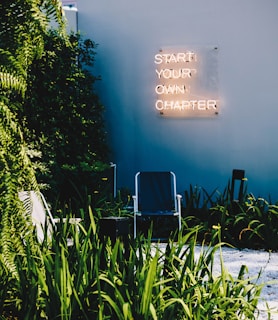 A garden area with a modern aesthetic features tall green plants and a small seating area with a simple chair set on gravel. A neon sign mounted on a wall displays the message 'Start Your Own Chapter' in glowing letters.