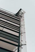 A modern building facade with large glass panels and metal framework, viewed from a low angle against a pale sky. The architectural design features clean lines and industrial elements, emphasizing transparency and structural integrity.