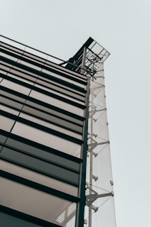 A modern building facade with large glass panels and metal framework, viewed from a low angle against a pale sky. The architectural design features clean lines and industrial elements, emphasizing transparency and structural integrity.