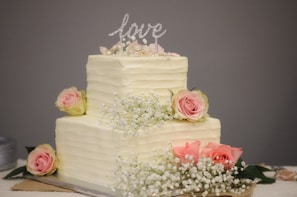 A two-tiered cake with textured white frosting, adorned with pink roses and delicate white baby's breath flowers. At the top, there is a glittery decoration spelling the word 'love'.