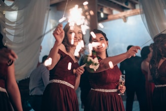Two women in elegant burgundy dresses are smiling and holding sparklers, surrounded by a softly lit reception area with draped fabric and a blue backdrop. One holds a bouquet and the other holds a champagne flute.