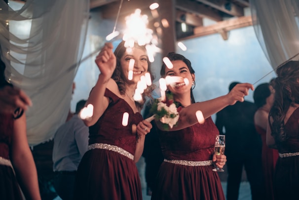 Two women in elegant burgundy dresses are smiling and holding sparklers, surrounded by a softly lit reception area with draped fabric and a blue backdrop. One holds a bouquet and the other holds a champagne flute.