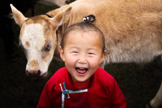 A young child wearing a bright red traditional outfit is smiling joyfully with an open mouth, seemingly in laughter, while standing beside a light brown and white calf with its fur slightly wet. The background appears to be grassy, creating a rustic, outdoor setting.