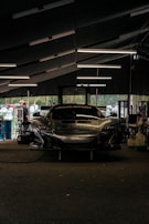 A sleek race car is parked inside a dimly-lit garage. The garage features modern overhead lighting, casting reflections on the glossy surface of the car. The front of the car displays a bold brand logo with a large air intake below it. Around the garage, various tools and equipment are scattered, and a few people are seen in the background, possibly mechanics or bystanders.