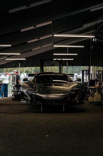 A sleek race car is parked inside a dimly-lit garage. The garage features modern overhead lighting, casting reflections on the glossy surface of the car. The front of the car displays a bold brand logo with a large air intake below it. Around the garage, various tools and equipment are scattered, and a few people are seen in the background, possibly mechanics or bystanders.