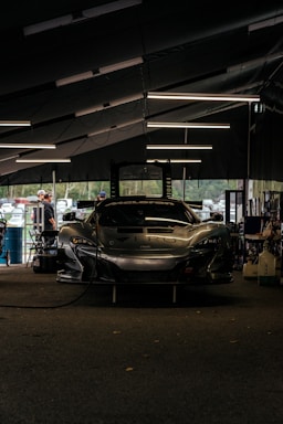 A sleek race car is parked inside a dimly-lit garage. The garage features modern overhead lighting, casting reflections on the glossy surface of the car. The front of the car displays a bold brand logo with a large air intake below it. Around the garage, various tools and equipment are scattered, and a few people are seen in the background, possibly mechanics or bystanders.