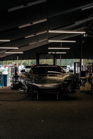 A sleek race car is parked inside a dimly-lit garage. The garage features modern overhead lighting, casting reflections on the glossy surface of the car. The front of the car displays a bold brand logo with a large air intake below it. Around the garage, various tools and equipment are scattered, and a few people are seen in the background, possibly mechanics or bystanders.