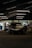 A sleek race car is parked inside a dimly-lit garage. The garage features modern overhead lighting, casting reflections on the glossy surface of the car. The front of the car displays a bold brand logo with a large air intake below it. Around the garage, various tools and equipment are scattered, and a few people are seen in the background, possibly mechanics or bystanders.