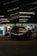 A sleek race car is parked inside a dimly-lit garage. The garage features modern overhead lighting, casting reflections on the glossy surface of the car. The front of the car displays a bold brand logo with a large air intake below it. Around the garage, various tools and equipment are scattered, and a few people are seen in the background, possibly mechanics or bystanders.