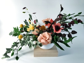A realistic arrangement of fabric or artificial flowers in various colors including peach, pink, yellow, and burgundy, set in a simple white vase on a wooden block. The flowers and leaves are detailed, showcasing a mix of roses, poppies, and other botanical elements with verdant green foliage.