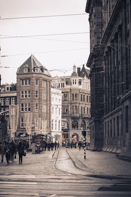 A European city street scene with historic architecture, featuring ornate buildings with decorative facades. The cobblestone street is bustling with pedestrians, and tram tracks run along the center. Shops and streetlights are visible, adding to the lively atmosphere.