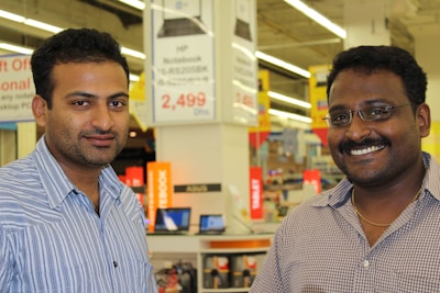 Two men are standing in a retail store, smiling at the camera. The background shows store signage and electronic products on display, including laptops.