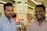 Two men are standing in a retail store, smiling at the camera. The background shows store signage and electronic products on display, including laptops.
