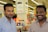 Two men are standing in a retail store, smiling at the camera. The background shows store signage and electronic products on display, including laptops.