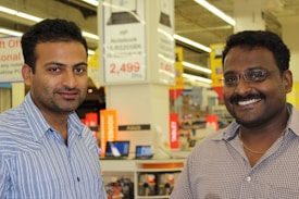Two men are standing in a retail store, smiling at the camera. The background shows store signage and electronic products on display, including laptops.