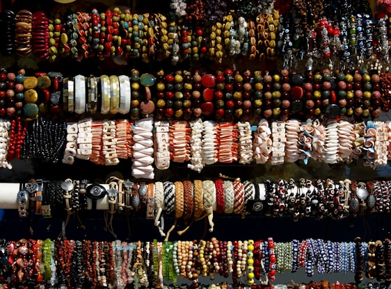 An assortment of colorful bracelets arranged in rows. The bracelets feature various designs and materials, including beads, shells, leather, and woven patterns. The display suggests a vibrant and diverse collection, showcasing a range of artistic styles.