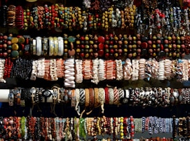 An assortment of colorful bracelets arranged in rows. The bracelets feature various designs and materials, including beads, shells, leather, and woven patterns. The display suggests a vibrant and diverse collection, showcasing a range of artistic styles.
