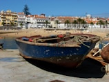 A small, weathered fishing boat sits on a concrete dock overlooking a sandy beach with several people sunbathing and enjoying the water. In the background, there are colorful buildings with red-tiled roofs and palm trees lining a promenade.