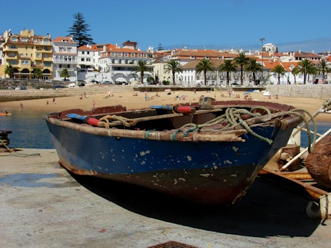 A small, weathered fishing boat sits on a concrete dock overlooking a sandy beach with several people sunbathing and enjoying the water. In the background, there are colorful buildings with red-tiled roofs and palm trees lining a promenade.