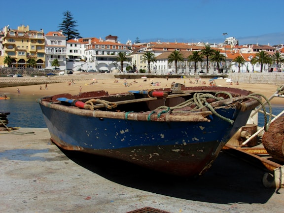 A small, weathered fishing boat sits on a concrete dock overlooking a sandy beach with several people sunbathing and enjoying the water. In the background, there are colorful buildings with red-tiled roofs and palm trees lining a promenade.