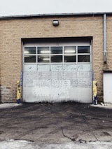 A large industrial garage door set into a tan brick building. The door is white with multiple small windows at the top, and there are yellow and blue protective bollards on either side. The ground in front of the door is paved and appears slightly worn.