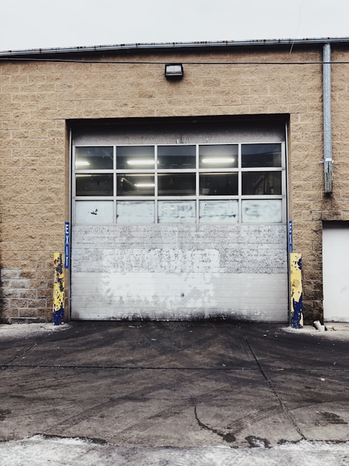 A large industrial garage door set into a tan brick building. The door is white with multiple small windows at the top, and there are yellow and blue protective bollards on either side. The ground in front of the door is paved and appears slightly worn.