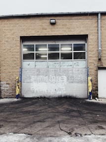 A large industrial garage door set into a tan brick building. The door is white with multiple small windows at the top, and there are yellow and blue protective bollards on either side. The ground in front of the door is paved and appears slightly worn.