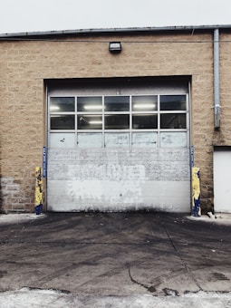 A large industrial garage door set into a tan brick building. The door is white with multiple small windows at the top, and there are yellow and blue protective bollards on either side. The ground in front of the door is paved and appears slightly worn.