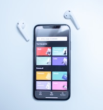A smartphone displaying a music streaming app interface is placed on a light blue surface. The screen shows various musical genres and options in colorful boxes, including Rock, Electronic/Dance, Pop, Indie, Podcasts, Charts, New Releases, and Radio. Two white wireless earbuds are positioned on either side of the phone.