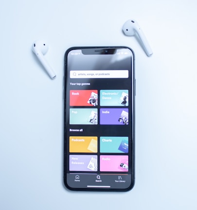 A smartphone displaying a music streaming app interface is placed on a light blue surface. The screen shows various musical genres and options in colorful boxes, including Rock, Electronic/Dance, Pop, Indie, Podcasts, Charts, New Releases, and Radio. Two white wireless earbuds are positioned on either side of the phone.