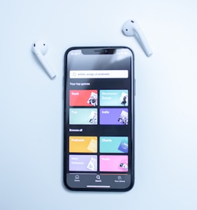 A smartphone displaying a music streaming app interface is placed on a light blue surface. The screen shows various musical genres and options in colorful boxes, including Rock, Electronic/Dance, Pop, Indie, Podcasts, Charts, New Releases, and Radio. Two white wireless earbuds are positioned on either side of the phone.