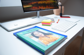 A modern office workspace with a white desk featuring a computer monitor displaying a landscape image, a white keyboard, and a closed laptop. In the foreground, a magazine titled 'Offscreen' with a cover image is prominently placed. Additional items include a stack of colorful sticky notes, a pen, and a decorative plant in a pot.