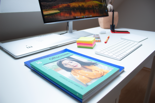 A modern office workspace with a white desk featuring a computer monitor displaying a landscape image, a white keyboard, and a closed laptop. In the foreground, a magazine titled 'Offscreen' with a cover image is prominently placed. Additional items include a stack of colorful sticky notes, a pen, and a decorative plant in a pot.