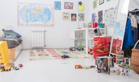 A cluttered children's bedroom with a bed, a colorful rug, and various toys and books scattered across the floor. A desk with a chair holds art supplies and a laptop. The walls are decorated with maps, posters, and artwork. A basket with clothes and a full-length mirror are visible, with coats hanging nearby.