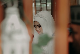 A woman wearing an intricately adorned hijab with a beaded headpiece, in a contemplative pose. The setting appears to be indoors, with blurred objects in the foreground contributing to a soft-focus effect. The color palette includes warm, muted tones.