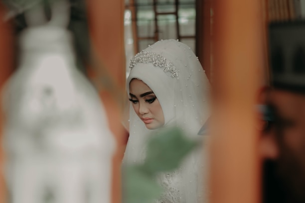 A woman wearing an intricately adorned hijab with a beaded headpiece, in a contemplative pose. The setting appears to be indoors, with blurred objects in the foreground contributing to a soft-focus effect. The color palette includes warm, muted tones.