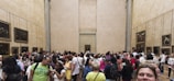 A large group of people gathered in a spacious gallery room with high beige walls adorned with framed paintings. It appears to be an art museum with a crowded and lively atmosphere, as visitors engage and take photos.