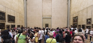 A large group of people gathered in a spacious gallery room with high beige walls adorned with framed paintings. It appears to be an art museum with a crowded and lively atmosphere, as visitors engage and take photos.