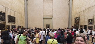 A large group of people gathered in a spacious gallery room with high beige walls adorned with framed paintings. It appears to be an art museum with a crowded and lively atmosphere, as visitors engage and take photos.