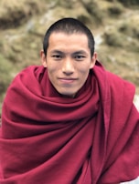 A person is wearing a deep red robe and has a serene expression with close-cropped hair. The background consists of blurred natural scenery with earthy tones.