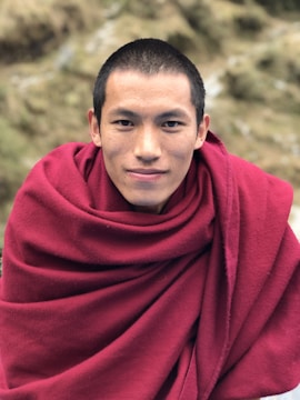 A person is wearing a deep red robe and has a serene expression with close-cropped hair. The background consists of blurred natural scenery with earthy tones.