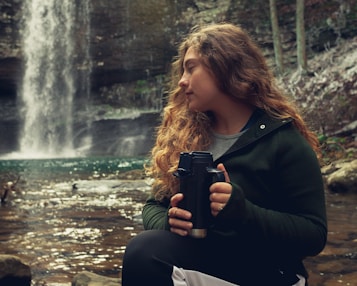 A person with long curly hair is sitting by a serene waterfall, holding a black travel mug. The surrounding environment is natural, with rocks and flowing water visible. The person appears to be relaxed, gazing thoughtfully into the distance.